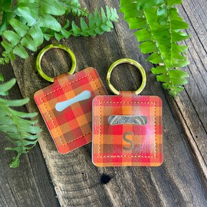 May include: Two red and orange plaid keyrings with gold rings. One keyring has a clear pocket and the other has a pocket with a coin inside. The coin has the letter "S" on it.