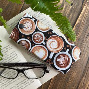 May include: A black eyeglasses case with a coffee-themed pattern featuring various latte art designs in shades of brown and white. The case is open, revealing a pair of eyeglasses resting on a book. A fern and wooden surface are in the background.