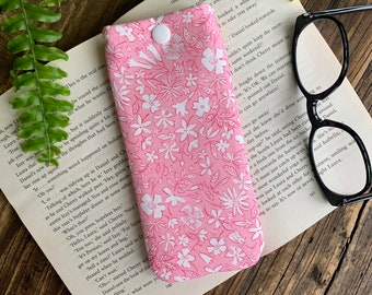 May include: A pink and white floral glasses case with a white button closure. The case is lying on a book with text.