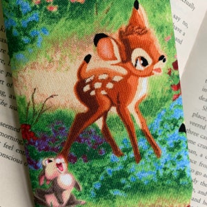 Puede incluir: Primer plano de una tela con una imagen de dibujos animados de Bambi el ciervo y Conejo. La tela es verde con flores azules y un fondo marr&oacute;n claro.