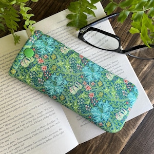 May include: A green and blue floral patterned glasses case with a green button closure. The case is resting on an open book with black-framed glasses nearby. The case is approximately 7 inches long.