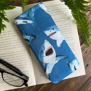 May include: A blue fabric glasses case featuring a pattern of cartoon sharks with open mouths and sharp teeth. The case has a button closure and is resting on an open book. The case is designed to protect eyeglasses.