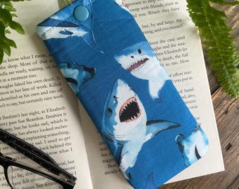 May include: A blue fabric glasses case featuring a pattern of cartoon sharks with open mouths and sharp teeth. The case has a button closure and is resting on an open book. The case is designed to protect eyeglasses.
