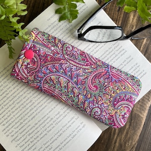 May include: A patterned glasses case in pink, purple, and blue, featuring a paisley design. It has a pink button closure and is placed on an open book, next to a pair of black glasses.