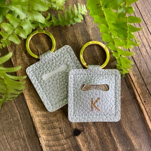 May include: Two silver leather keyrings with gold rings. One keyring has a pocket for a coin and the letter "K" embossed on the front. The other keyring has a blank pocket.