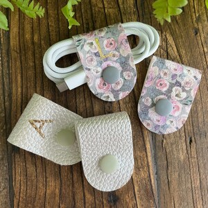 May include: Three cable organizers in different styles. One is white with a floral pattern and the letter "L" embroidered on it. Another is white with a floral pattern. The third is plain white with the letter "A" embroidered on it.