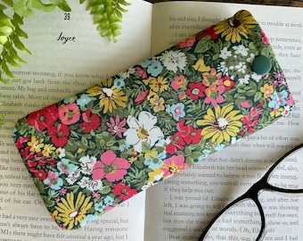 May include: A floral spectacles case with a green button fastening. The case has a dark green background with a pattern of yellow, red, pink, and white flowers. The case is on an open book.