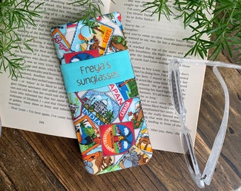 May include: A blue and white fabric sunglasses case with a colorful collage of travel stamps and the text "Freya's sunglasses" printed on the front.