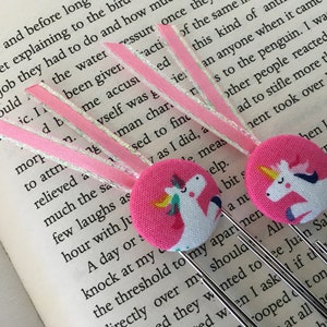 May include: Two pink fabric unicorn bookmarks with silver metal clips. The unicorns are white with rainbow manes and tails. The bookmarks are on a page of a book.