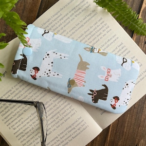May include: A light blue glasses case featuring a repeating pattern of cartoon dogs wearing glasses and sweaters. The case has a button closure and is on an open book.