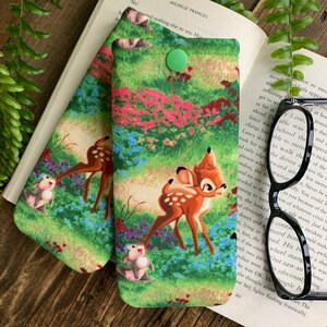 Puede incluir: Dos estuches para gafas de tela verde y marr&oacute;n con cierre de bot&oacute;n. Los estuches presentan un patr&oacute;n repetido de un ciervo y un conejo de dibujos animados en un entorno de bosque.