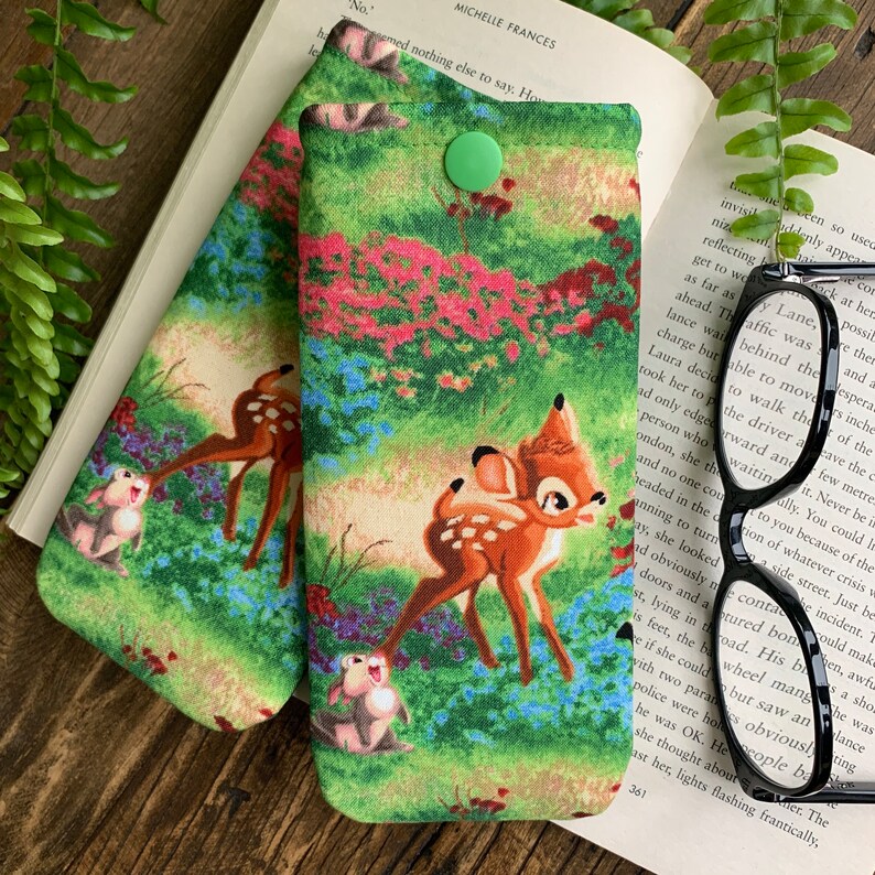 Puede incluir: Dos estuches para gafas de tela verde y marr&oacute;n con cierre de bot&oacute;n. Los estuches presentan un patr&oacute;n repetido de un ciervo y un conejo de dibujos animados en un entorno de bosque.