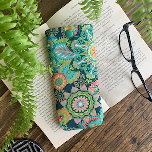 May include: A patterned glasses case in teal, green, and orange floral fabric. The case is closed with a green button and sits on an open book. A pair of black glasses is next to the case.