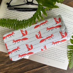 Peut inclure: Un étui à lunettes blanc avec un motif répété de renards rouges et de lignes pointillées noires. L'étui est fermé par un bouton blanc. L'étui est posé sur un livre ouvert.