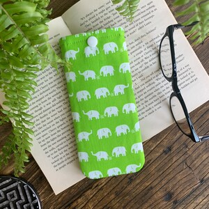 Peut inclure: Un étui à lunettes en tissu vert avec une fermeture à bouton blanc et un motif répété d'éléphants blancs sur fond vert.