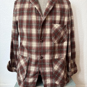 May include: A long-sleeve blazer featuring a plaid pattern in shades of dark red, cream, and muted green. The garment includes a notched lapel, a single chest pocket, two large lower patch pockets, and two dark brown buttons on the front closure.