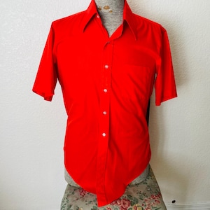 Op de afbeelding: Een felrood shirt met korte mouwen, een klassieke kraag en een enkele borstzak. Het shirt is dichtgeknoopt met witte knopen en wordt op een mannequin gepresenteerd. De stof van het shirt lijkt glad en kreukvrij.