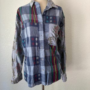 Può includere: Una camicia a maniche lunghe con bottoni caratterizzata da un design patchwork con quadri blu, motivi floreali e strisce verticali rosse, gialle e verdi. La camicia presenta un colletto, una tasca sul petto e bottoni chiari sulla parte anteriore.