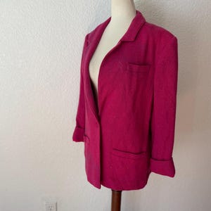 May include: A magenta blazer with a notched lapel, two front pockets, and folded cuffs. The blazer is displayed on a mannequin. The garment has a textured appearance and a classic, tailored design, suitable for various occasions.