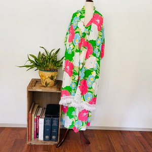 May include: A green, white, and pink floral print maxi dress with white fringe trim. The dress is on a mannequin and is displayed against a white wall.