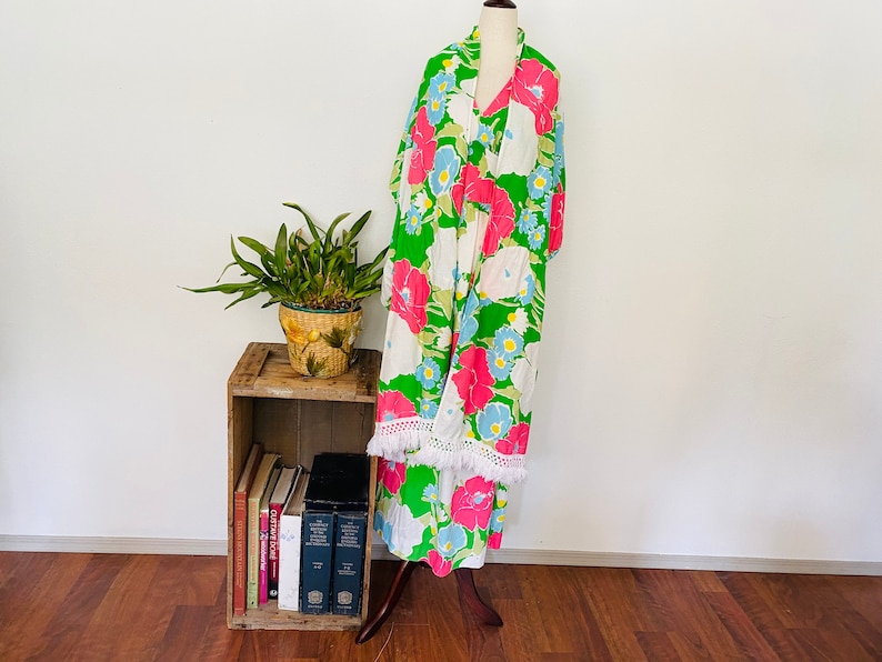 May include: A green, white, and pink floral print maxi dress with white fringe trim. The dress is on a mannequin and is displayed against a white wall.