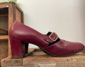 May include: A pair of burgundy leather heeled shoes with a buckle strap. The shoes have a chunky heel and a pointed toe.