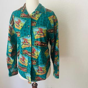 May include: A teal button-down shirt with a repeating pattern of cowboy illustrations and the words "OUT WEST". The long-sleeved shirt has a collared neckline and is displayed on a mannequin.