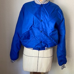 K&ouml;nnte beinhalten: Eine blaue Satin-Bomberjacke mit einem wei&szlig;-blau gestreiften Kragen und B&uuml;ndchen. Die Jacke hat einen Druckknopfverschluss und zwei Taschen.