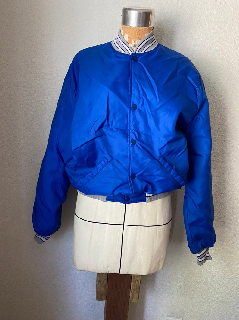K&ouml;nnte beinhalten: Eine blaue Satin-Bomberjacke mit einem wei&szlig;-blau gestreiften Kragen und B&uuml;ndchen. Die Jacke hat einen Druckknopfverschluss und zwei Taschen.