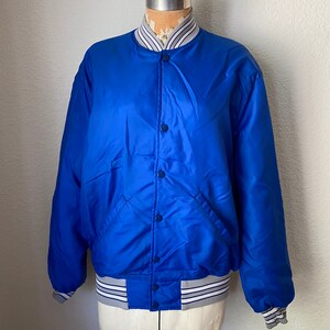 K&ouml;nnte beinhalten: Eine blaue Satin-Bomberjacke mit wei&szlig;-blau gestreiftem Besatz an Kragen, &Auml;rmelb&uuml;ndchen und Bund. Die Jacke hat einen Knopfverschluss und zwei Taschen.