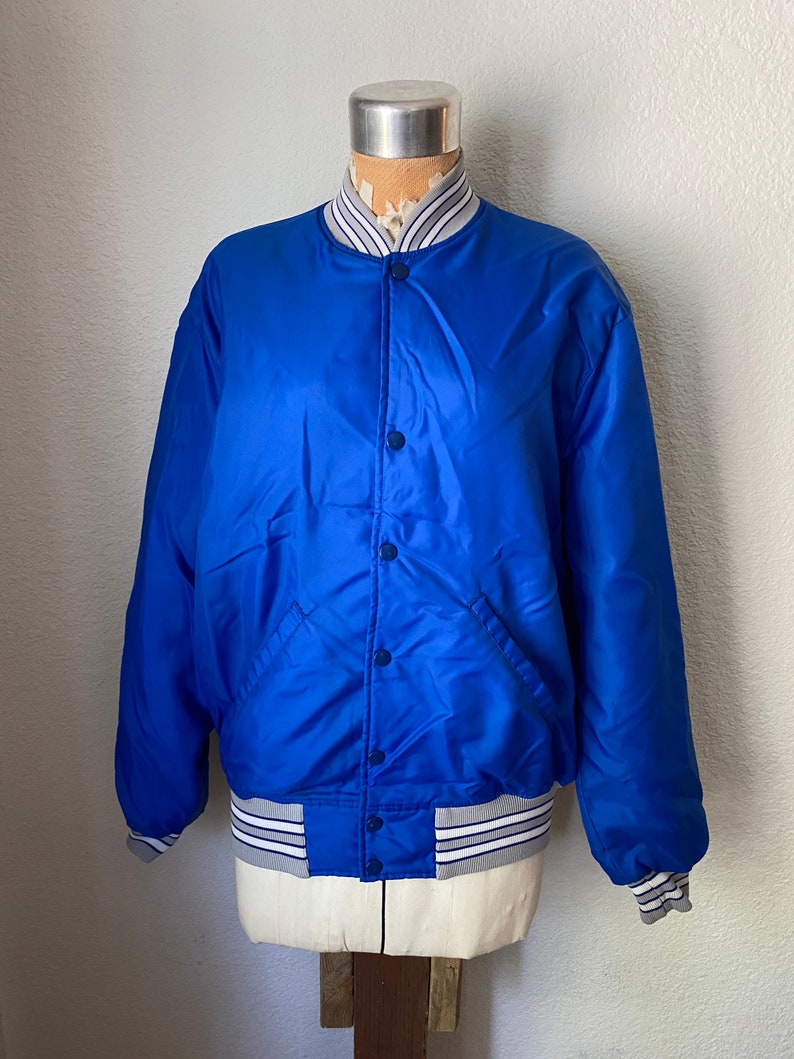 K&ouml;nnte beinhalten: Eine blaue Satin-Bomberjacke mit wei&szlig;-blau gestreiftem Besatz an Kragen, &Auml;rmelb&uuml;ndchen und Bund. Die Jacke hat einen Knopfverschluss und zwei Taschen.