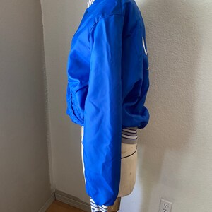 K&ouml;nnte beinhalten: Eine blaue Satin-Bomberjacke mit wei&szlig;en und grauen Streifen an den S&auml;umen. Die Jacke hat einen Rei&szlig;verschluss und ein wei&szlig;es Logo auf der R&uuml;ckseite.