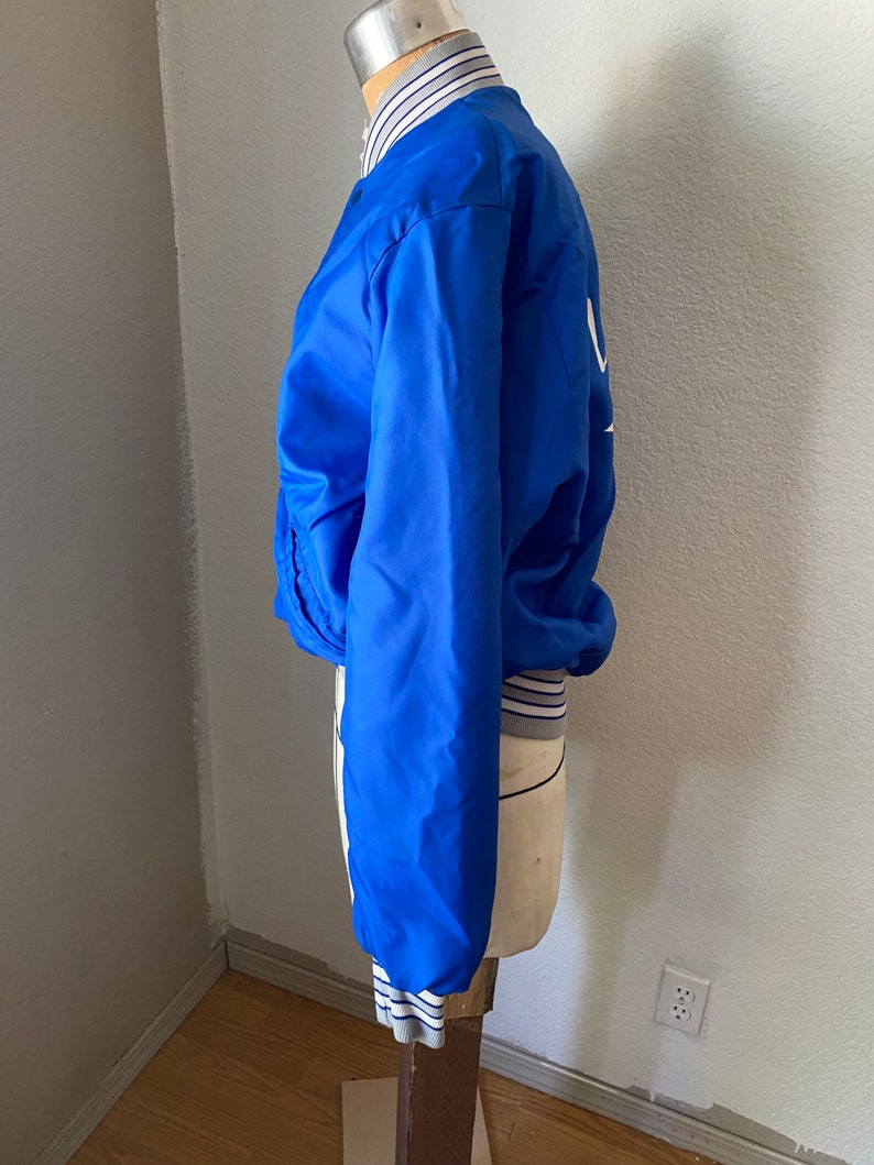 K&ouml;nnte beinhalten: Eine blaue Satin-Bomberjacke mit wei&szlig;en und grauen Streifen an den S&auml;umen. Die Jacke hat einen Rei&szlig;verschluss und ein wei&szlig;es Logo auf der R&uuml;ckseite.