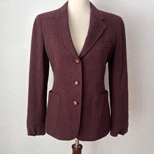 May include: A burgundy blazer with a two-button closure, a notched lapel, and two front pockets. The blazer is made of a textured fabric and has a classic, tailored design. The blazer is displayed on a mannequin.
