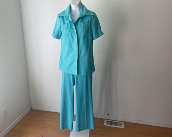 May include: A vintage turquoise two-piece pantsuit. The short-sleeved button-down shirt has two chest pockets and a collar. The pants are wide-legged. The outfit is displayed on a mannequin against a white wall.