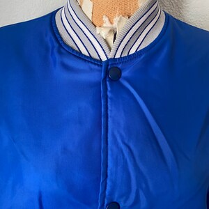 K&ouml;nnte beinhalten: Eine blaue Satin-Bomberjacke mit einem wei&szlig;-grauen gestreiften Kragen. Die Jacke hat einen Druckknopfverschluss und zwei Taschen.