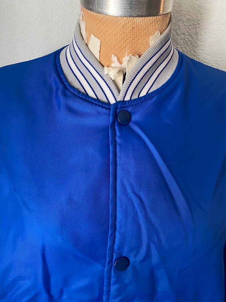 K&ouml;nnte beinhalten: Eine blaue Satin-Bomberjacke mit einem wei&szlig;-grauen gestreiften Kragen. Die Jacke hat einen Druckknopfverschluss und zwei Taschen.