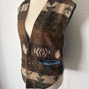 May include: A sleeveless vest featuring a woven brown, black, and tan geometric pattern on the front. The garment has a V-neckline, three metallic buttons down the center, and a single pocket with a light blue denim trim on the lower side.