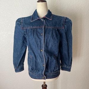 May include: A dark blue denim jacket with a collared neckline and button-up closure. The jacket features a red stitched trim and gathered detailing on the shoulders and waist. The jacket is displayed on a mannequin.