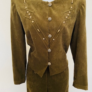 May include: A brown suede pantsuit with a cropped jacket. The jacket has a round neckline, long sleeves, and a button closure. The jacket is decorated with gold-toned buttons and studs in a geometric pattern. The pants have a straight leg and a high waist.