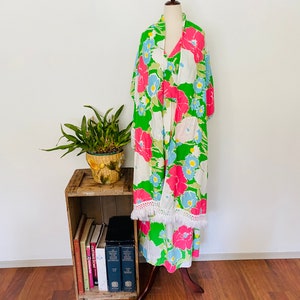 May include: A green, white, pink, and blue floral print maxi dress with a white fringe trim. The dress is on a mannequin and is displayed on a wooden shelf with books.
