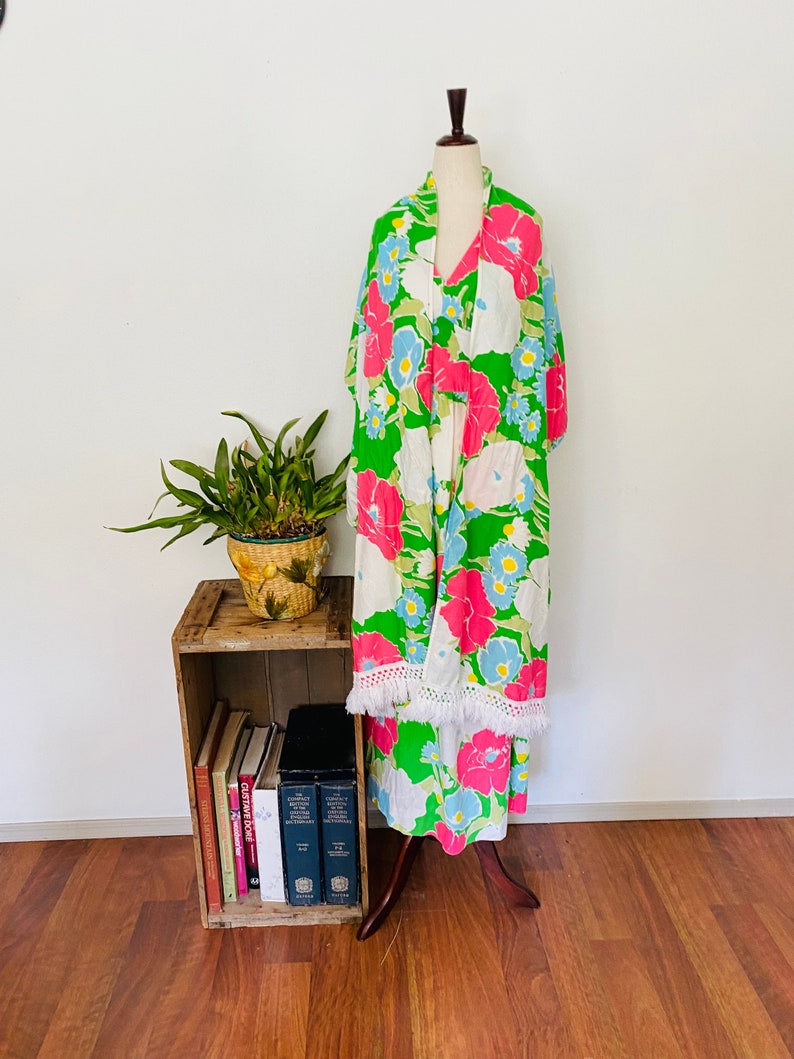 May include: A green, white, pink, and blue floral print maxi dress with a white fringe trim. The dress is on a mannequin and is displayed on a wooden shelf with books.