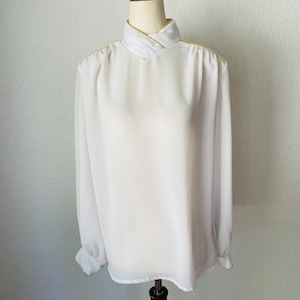 May include: A white blouse with long sleeves and a high collar, displayed on a mannequin. The collar and cuffs are trimmed with gold-colored accents. The blouse has a flowing, lightweight appearance, suggesting a vintage or retro style.
