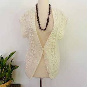 May include: Cream-coloured, open-front knitted vest with a textured pattern and short sleeves. The vest is displayed on a mannequin with a dark brown beaded necklace. The style is a classic, open cardigan.