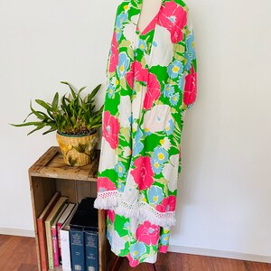 May include: A green, white, pink, and blue floral print maxi dress with a white crocheted trim. The dress is on a mannequin and is displayed on a wooden stand.