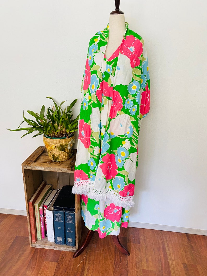 May include: A green, white, pink, and blue floral print maxi dress with a white crocheted trim. The dress is on a mannequin and is displayed on a wooden stand.