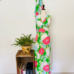 May include: A white maxi dress with a green, pink, and blue floral print. The dress has a V-neckline and sleeveless design. It is displayed on a mannequin against a white wall.