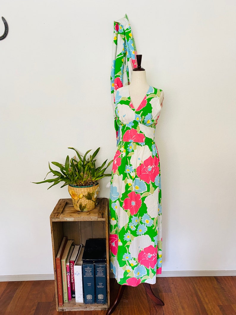 May include: A white maxi dress with a green, pink, and blue floral print. The dress has a V-neckline and sleeveless design. It is displayed on a mannequin against a white wall.