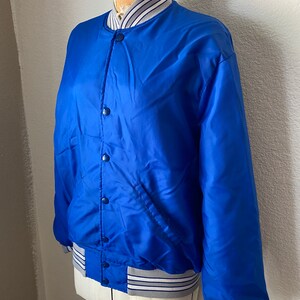 K&ouml;nnte beinhalten: Eine k&ouml;nigsblaue Satin-Bomberjacke mit einem wei&szlig;en und blauen gerippten Kragen, B&uuml;ndchen und Bund. Die Jacke hat einen Knopfverschluss und eine einzelne Tasche auf der linken Seite.