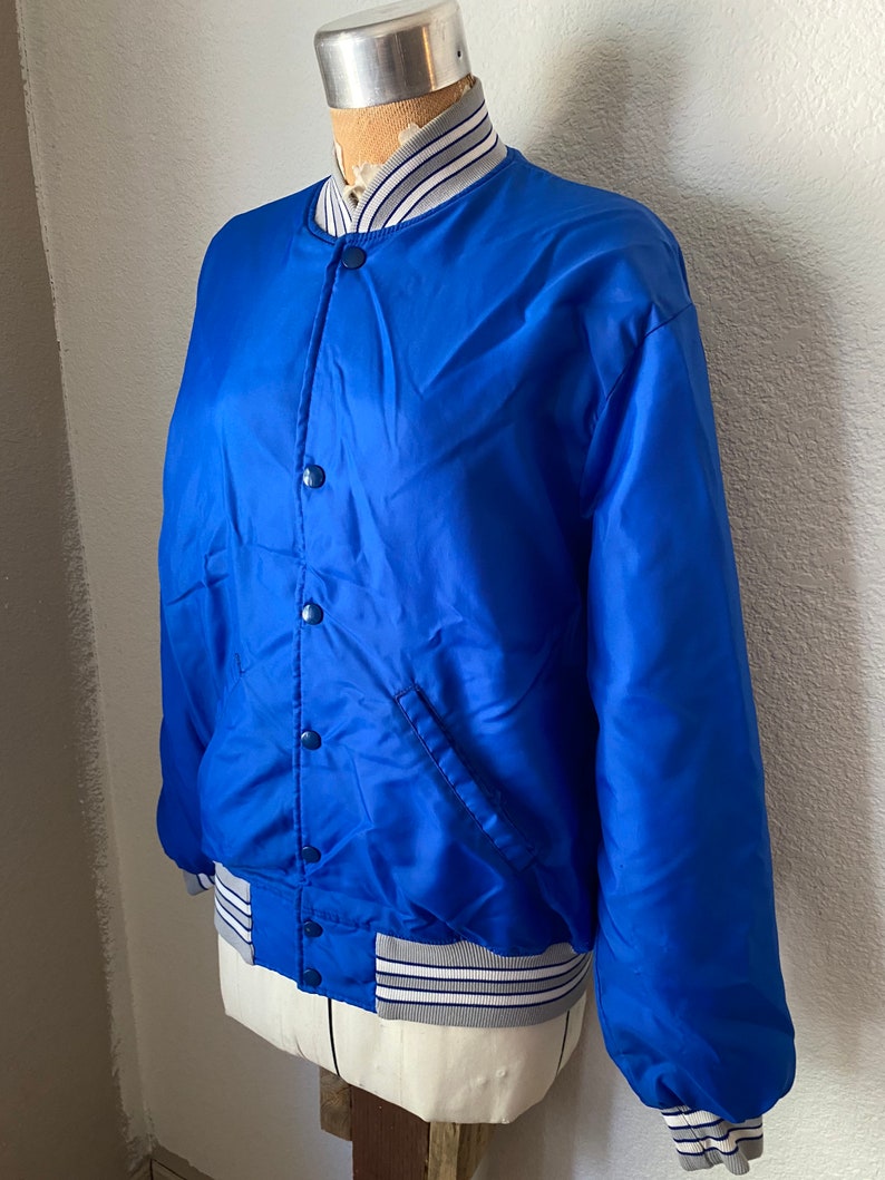 K&ouml;nnte beinhalten: Eine k&ouml;nigsblaue Satin-Bomberjacke mit einem wei&szlig;en und blauen gerippten Kragen, B&uuml;ndchen und Bund. Die Jacke hat einen Knopfverschluss und eine einzelne Tasche auf der linken Seite.